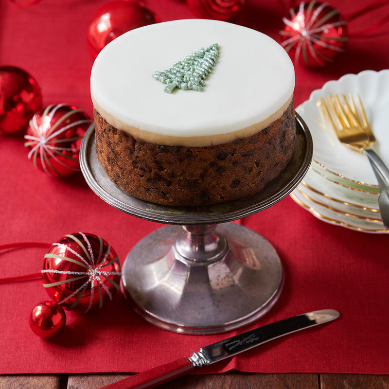 Christmas cake with white frosting and a tree decoration on a red tablecloth with festive ornaments.