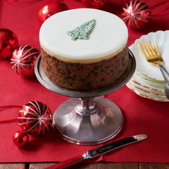 Christmas cake with white frosting and a tree decoration on a red tablecloth with festive ornaments. !